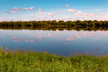 Summer sky with pink clouds reflecting in the water. Meadow and forest on the banks. Summer concept.