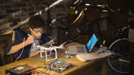 Student is soldering electrical components on a drone in a garage while checking a laptop computer.