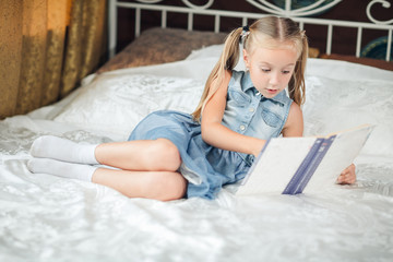 Cute little girl in denim sundress reading book on bed at home