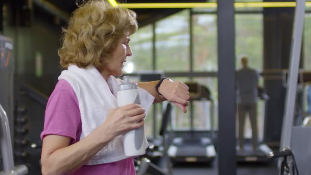 Medium shot of old Caucasian woman standing with towel on her shoulders, drinking water from sport bottle and checking her smart watch