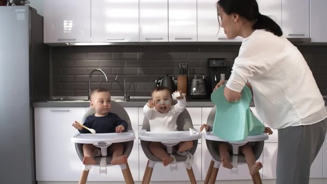 Full shot of happy 1-year-old Asian triplets, dressed in bodysuits, sitting together in high chairs in kitchen at home, and mother putting plastic feeding bibs on their necks
