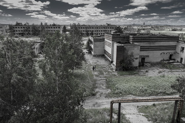 Landscape of abandoned city, ghost town, with modern, but rusty and ruined empty houses, barracks, and two lonely little figures of rambler adventurers inside. Post apocalypse world
