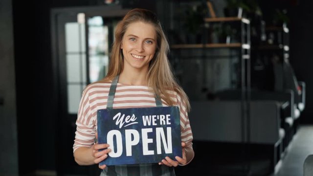 Portrait of attractive blond waitress holding open sign in new cafe smiling standing alone looking at camera with happy face. Modern youth and business concept.