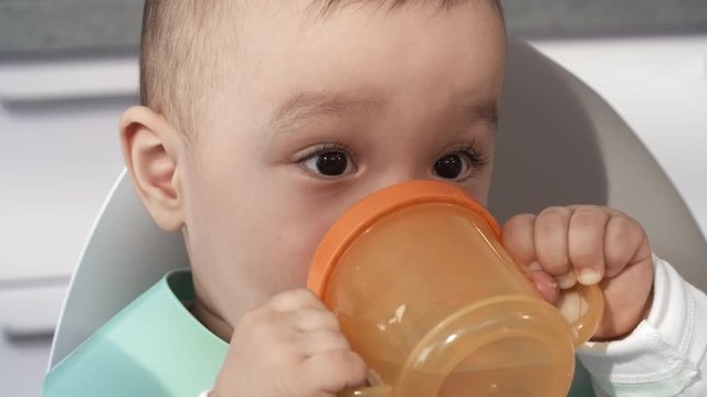 Close-up face shot of sloppy 1-year-old Asian baby with beautiful dark brown eyes, wearing plastic bib, sitting in high chair and drinking from orange plastic beaker cup, holding it with both hands