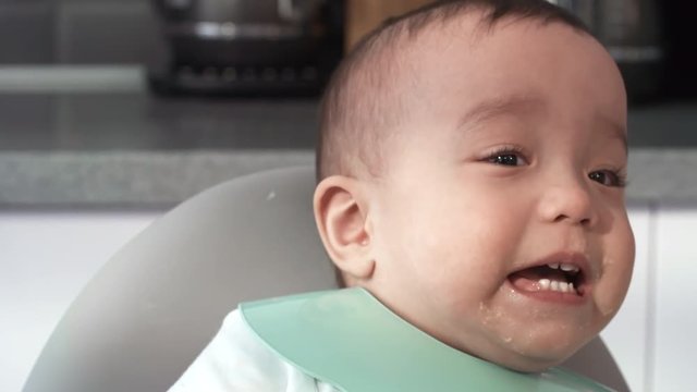 Close-up face shot of young Filipino baby with puree around mouth and beautiful brown eyes, wearing bright plastic bib, waving arms to attract attention, whimpering and appearing upset