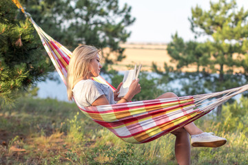 young attractive caucasian woman reading book in hammock in the forest, lake on the background