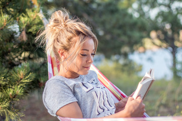 young attractive caucasian woman reading book in hammock in the forest, lake on the background