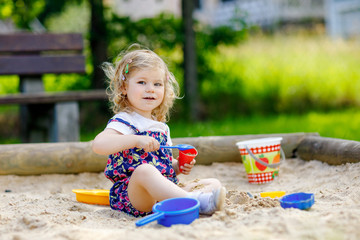 Cute toddler girl playing in sand on outdoor playground. Beautiful baby in red trousers having fun on sunny warm summer day. Child with colorful sand toys. Healthy active baby outdoors plays games