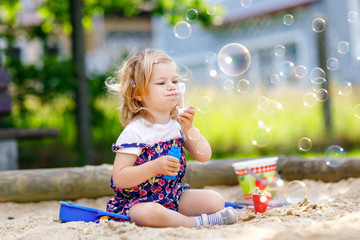 Beautiful little blonde toddler girl having fun with blowing soap bubble blower. Cute adorable baby...