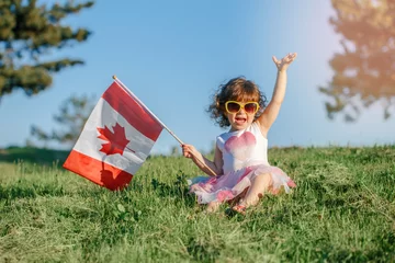 Fotobehang Portret van een schattig klein blank peutermeisje dat op groen gras in het park buiten zit en een grote Canadese vlag vasthoudt. Kid kind burger viert Canada Day op 1 juli. © anoushkatoronto