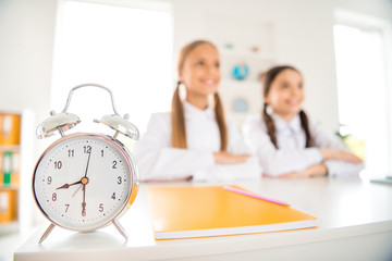 Close-up view of alarm clock two person nice attractive smart clever diligent cheerful cheery girls sitting still in modern light white interior style class room indoors