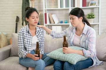 Best friends trying to comfort and cheer up young depressed woman after break up sitting on sofa at home. two women drink bottle beer on couch. sister touching girl shoulder consoling calm down