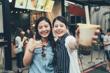 Naklejka premium two pretty asian women tourist travel in taiwan taipei holding modern asian beverage bubble milk tea. girls showing face camera with tasty drink local specialty smiling with thumb up good on street