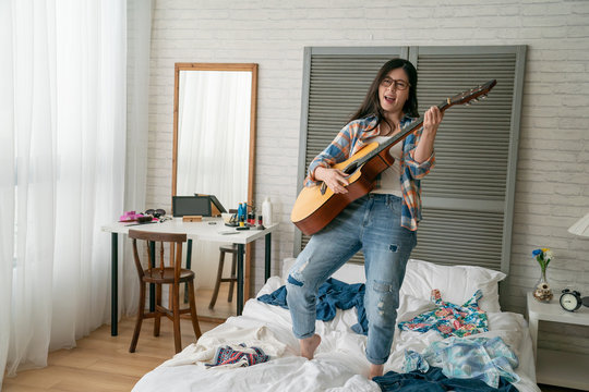 Asian Korean Teenage Girl Jumping Whilst Playing Guitar On Messy Bed With Clothes. Crazy Happy Young Woman In Eyeglasses Having Fun With Instrument Love Music Singing Songs At Home In Day Time.