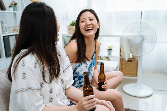 Happy Friends Talking Laughing Drinking Ice Bottle Beer Sitting On Sofa Cheerful. Young Girl Friends In Living Room Relax At Home With Electric Fan. Joy Two Ladies Enjoy Air Cooler Blowing Hair.