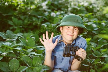 Portrait of little explorer with binoculars in forest. Boy traveler in helmet play in the park. Happy child go hiking with backpack in summer nature.