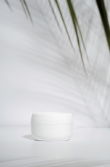White jar of cream on a white background with tropical palm leaves and their shadow. Stylish look of the product, mock up, identity.
