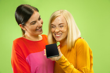 Portrait of pretty charming girls in casual outfits isolated on green studio background. Girlfriends or lesbians talking on smartphone. Concept of LGBT, equality, human emotions, love, relation.