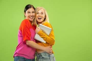 Portrait of pretty charming girls in casual outfits isolated on green studio background. Girlfriends or lesbians using a tablet for fun or payments. Concept of LGBT, human emotions, love, relation.