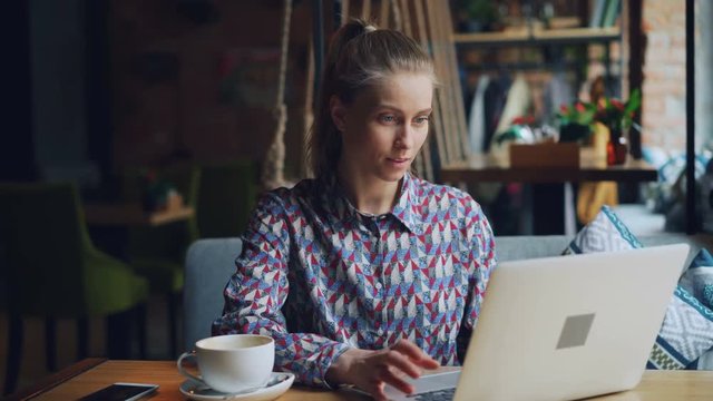 Happy young lady working with laptop in cafe typing sitting at table alone looking at computer screen concentrated on work. Modern technology and youth concept.