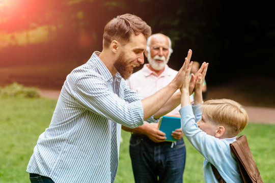 Image of a modern beard father giving a hi-five his little son with grandfather on background in the park. Dad and granddad meets their son from elementary school. the end of the school day concept