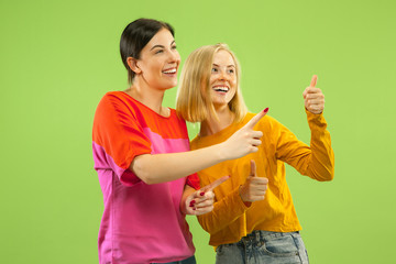 Portrait of pretty charming girls in casual outfits isolated on green studio background. Two female models as a girlfriends or lesbians. Concept of LGBT, equality, human emotions, love, relation.