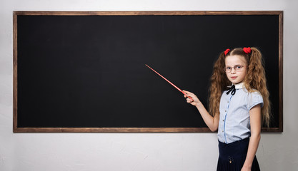 A girl schoolgirl is standing at the school board and holding a pointer.