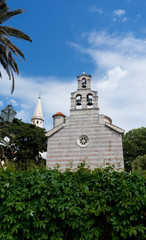 Obraz premium The Old Town of Budva, Montenegro - an old stone church adhering to the fortress of Budva to the Adriatic Sea. Flowers and palms in the city. Bells in a tower