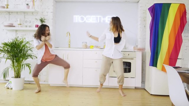 Women have fun in modern apartment. Side view crazy dancing girlfriend on kitchen with hanging rainbow flag. Girl making funny gesture,laughing and enjoying togetherness . Concept lgbt couple