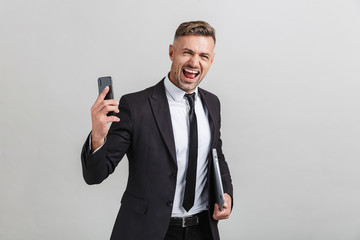 Portrait of ecstatic adult businessman in office suit holding smartphone and laptop while standing