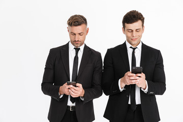 Image of successful serious two businessmen in office suits holding and typing on cellphones