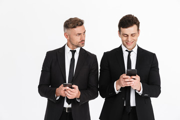 Image of handsome caucasian two businessmen in office suits holding and typing on cellphones
