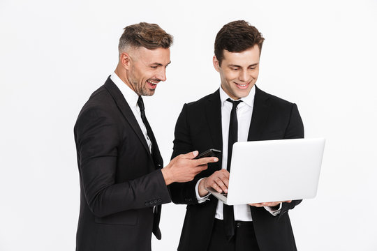 Image of two happy caucasian businessmen in office suits holding laptop and cellphones