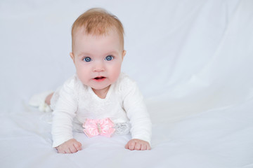 Cute baby with blue eyes in white dress with pink bow lying on her stomach looking at camera, white background,