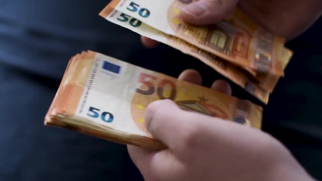 A steady close-up shot of hands counting money. Each fifty euro bill is carefully slid from one hand to another with a black, messy table-top cloth in the background. The stack is around 2000k euros.