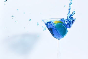 Cocktail with blue liquid in glass. Glass with blue water pouring with liquid with splashes and drops. Martini glass filling with alcohol with splashes on white background. Refreshing drink concept.