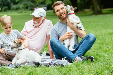 Grandfather, father and son sits, hugs, caress their two dogs Jack russel terrier on green grass in the summer park. Family Vacation. Generational connection. Pet, love friendship and people concept