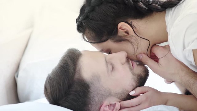 happy bearded man kissing cheeks and nose of attractive brunette girl in bedroom 