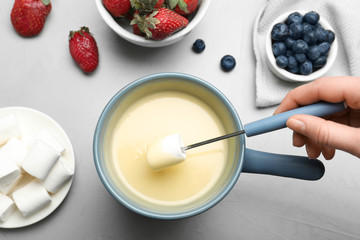 Woman dipping marshmallow into pot with white chocolate fondue at table, top view