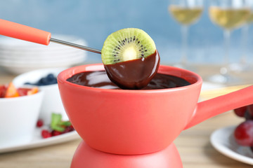 Dipping kiwi slice into pot with chocolate fondue on table, closeup