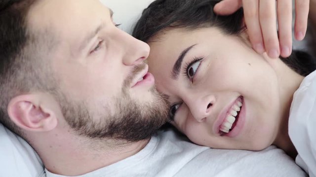 close up of happy man touching face and hair of cheerful woman while kissing and lying on bed 
