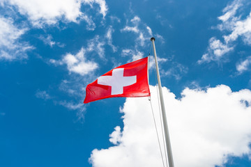 Waving in the wind to the left, the flag of Switzerland hanging on the mast, against the background of blue sky with white clouds.