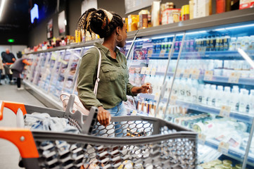 African woman with shopping cart choose yogurt bottle from fridge at supermarket.