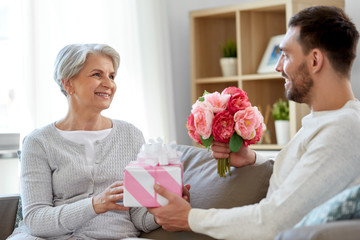 family, mother's day and birthday concept - smiling adult son giving present and flowers to his...