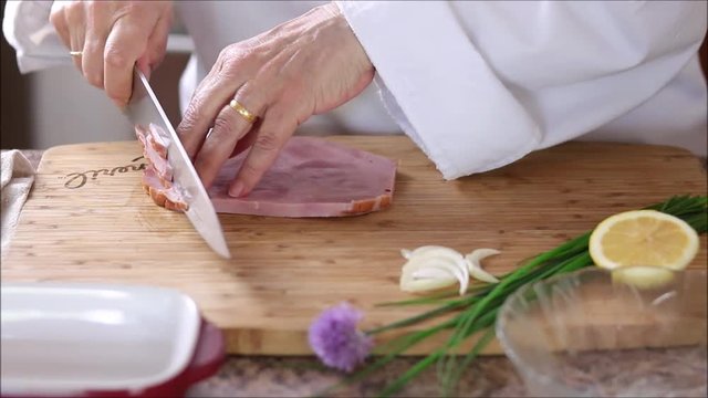 Chef in kitchen slicing rind off of thick cooked ham steak.