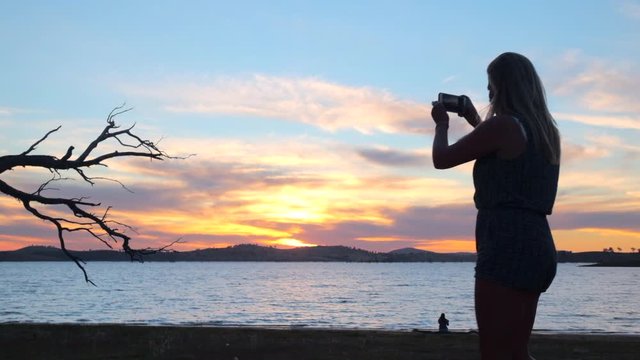 Tracking, slow motion shot following a woman walking and taking pictures, on her phone, of a tree at the beach, at lake Huma, on a sunny evening sunset, Hume Reservoir, New South Wales, in Australia