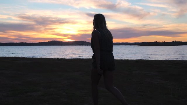 Tracking, slow motion shot of a woman walking on a beach, sunset in the background, at lake Huma, on a sunny evening, at Hume Reservoir, New South Wales, Australia