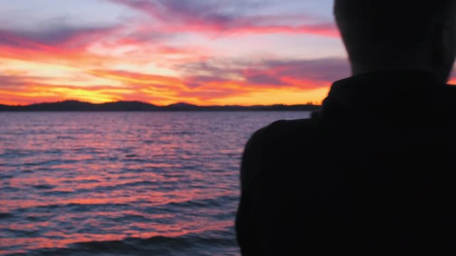 Dolly, slow motion shot of a man standing on a beach, looking at the sunset, at lake Huma, on a sunny evening dusk, at Hume Reservoir, New South Wales, Australia