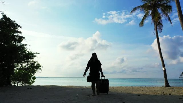 A woman puling a trolley bag starts to walk from a beach towards the camera. Next to palm trees on an island in bali. Blue Sky. Dolly out.
