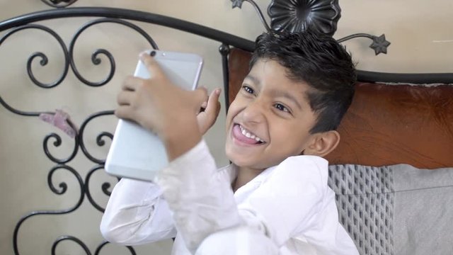 Cute adorable little indian asian caucasian boy child taking funny selfies on mobile phone or smart phone camera with funny expression in a bedroom close up shot side view
